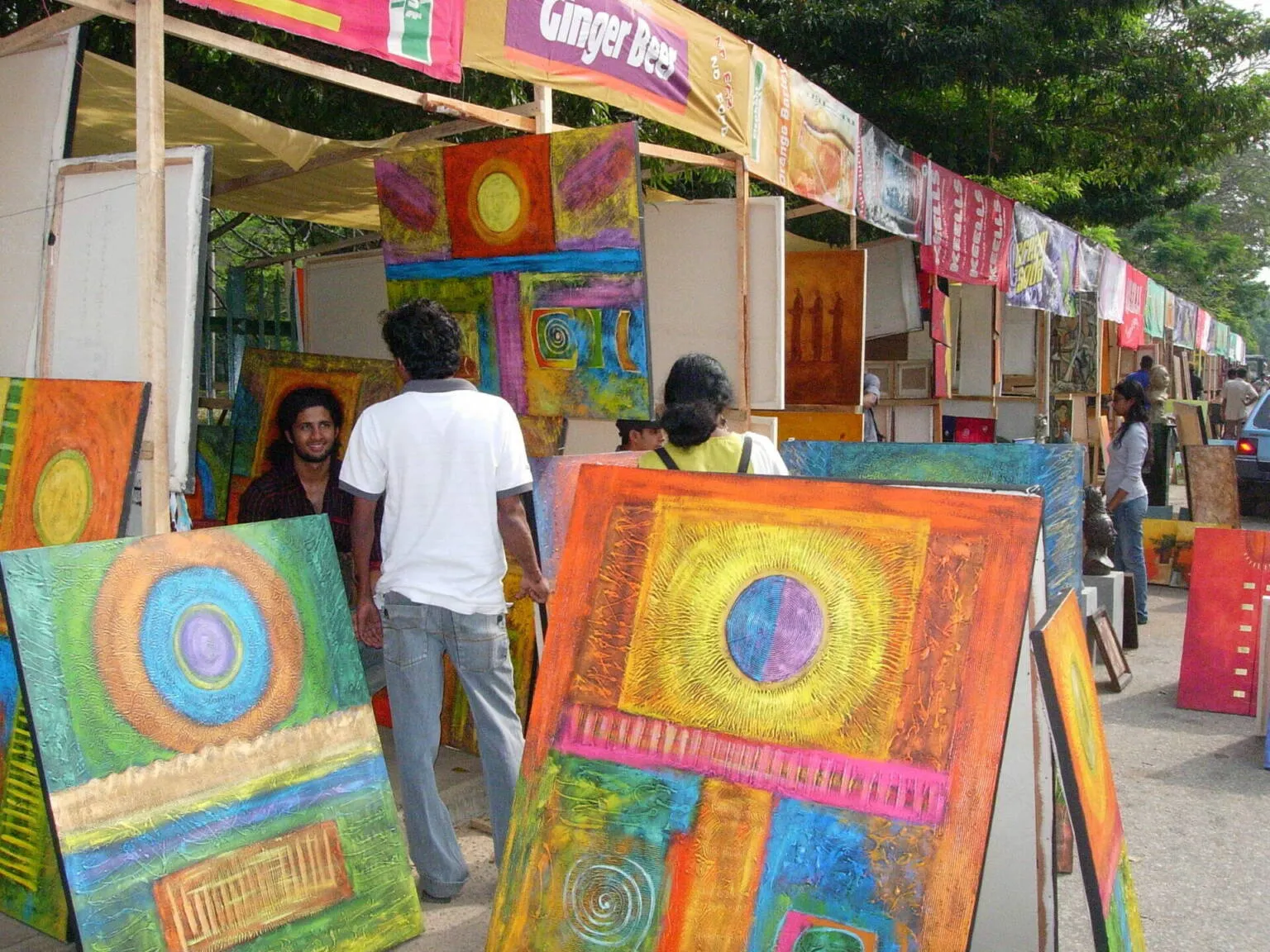 A diverse group of people admiring various paintings displayed at an outdoor market, surrounded by vibrant colors and art.